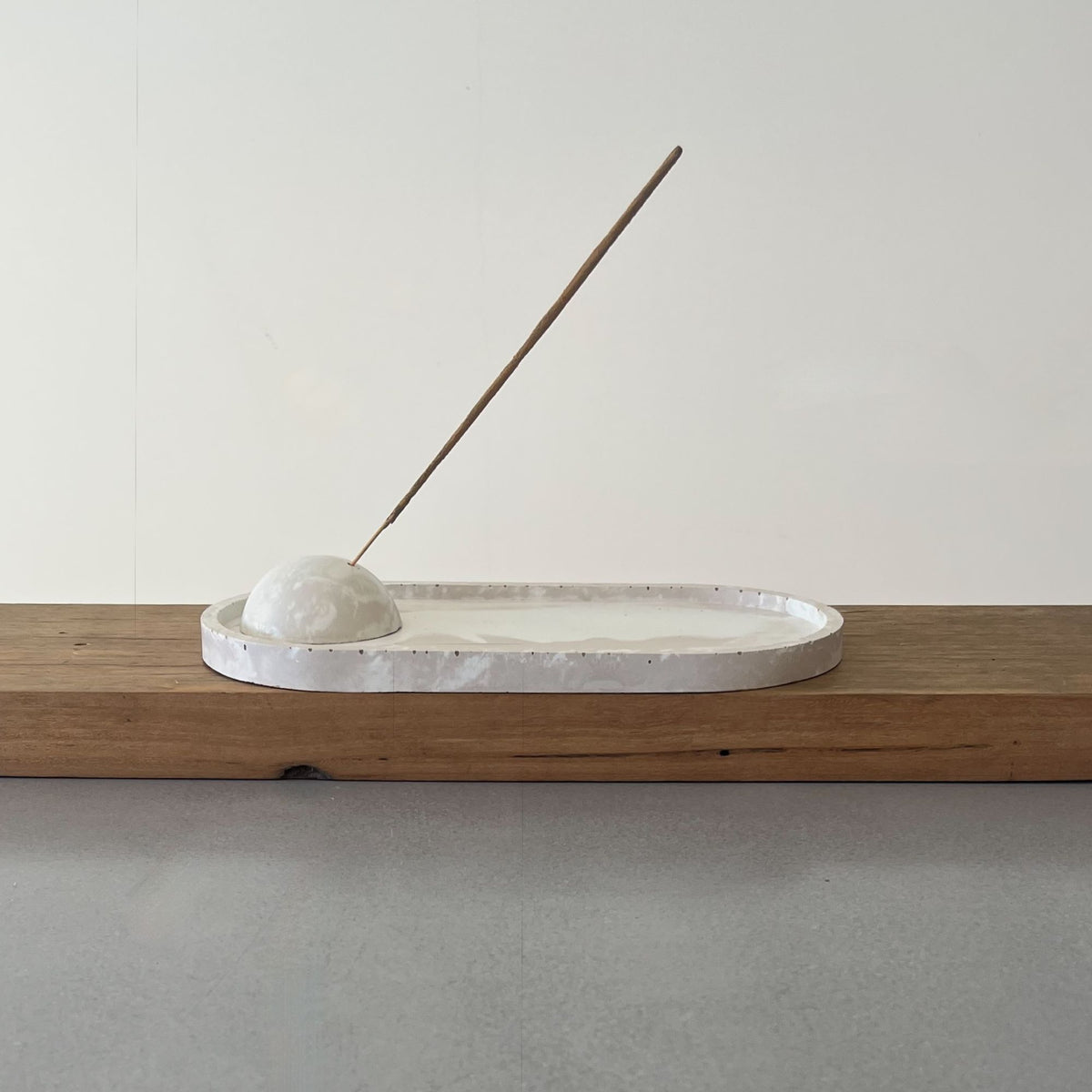 A minimalist white ceramic tray with a textured rim, holding a small white stone dish and a thin wooden incense stick, displayed on a wooden shelf against a neutral off-white wall.