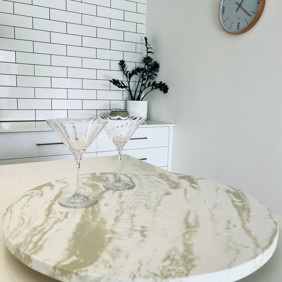 Lennoelia Designs 500 mm concrete Lazy Susan in Signature Marble finish styled with crystal martini glasses on a modern kitchen island. round white and beige marble-patterned countertop in a bright kitchen. White subway tile backsplash, white cabinets, and a wall clock are visible in the background.
