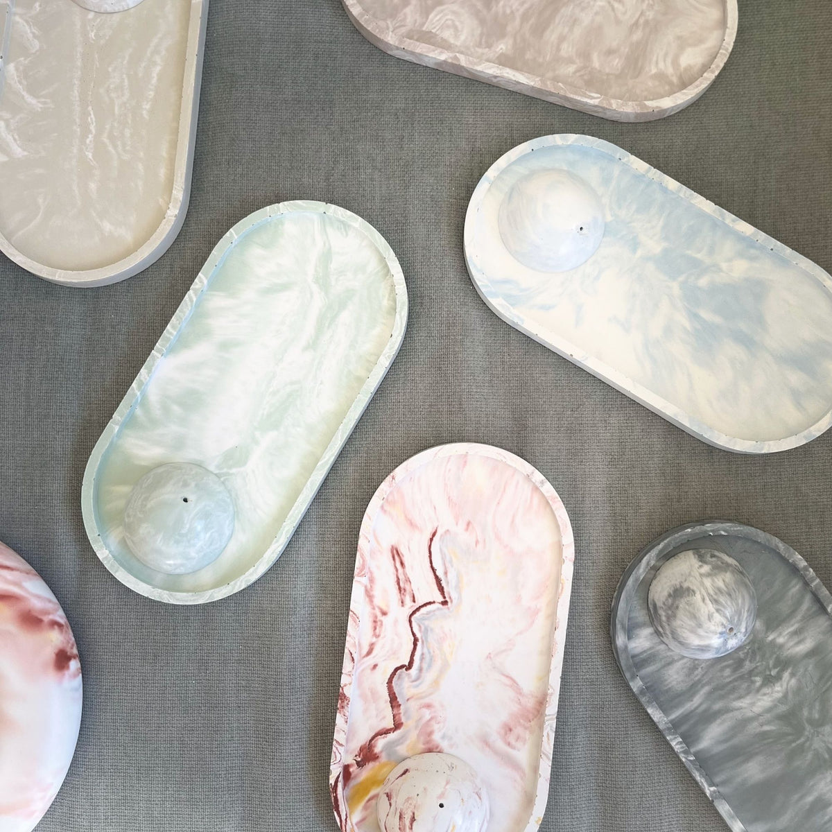 Several oval marble-style trays with small raised domes are arranged on a gray fabric background. The trays show different marbled colorways including cream, pale aqua, pale blue, pink, gray, and white swirls.