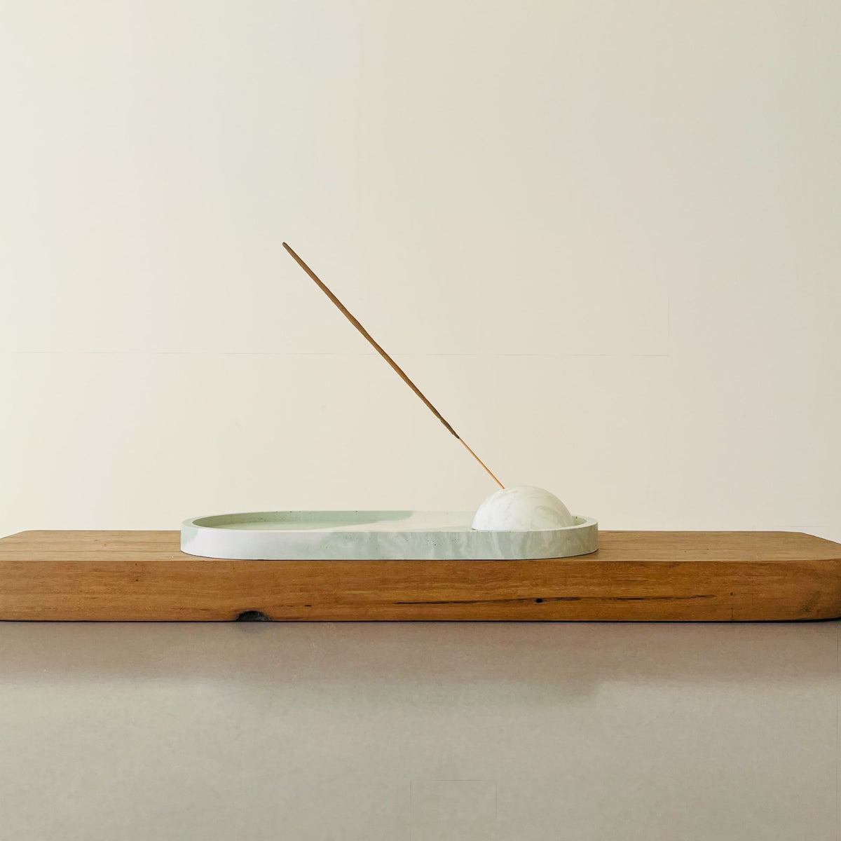 A minimalist incense holder with a Sea Meadow marble sphere and a shallow oval dish, set on a wooden block against a warm off-white background, with a single incense stick leaning diagonally.