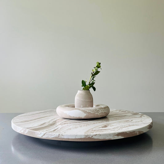 A round white and beige marble serving board on a gray table, styled with a small ribbed ceramic vase holding a green sprig, set against a neutral light gray wall.