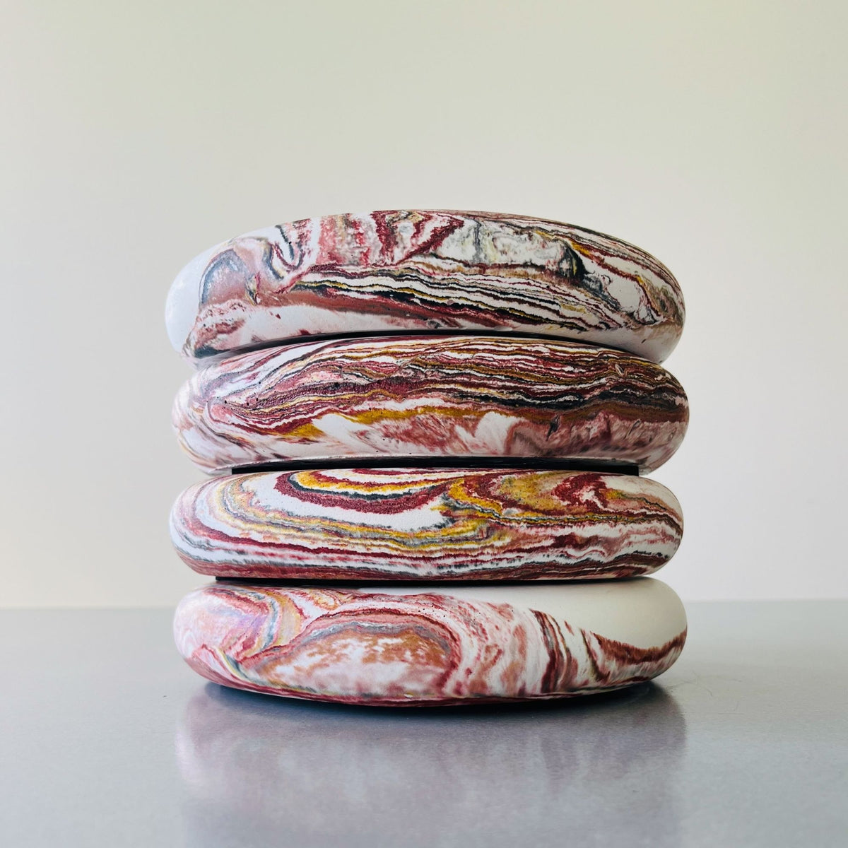 Four stacked bowls with a marbled pattern of red, white, yellow, and brown swirls, set on a reflective gray surface against a light beige background.