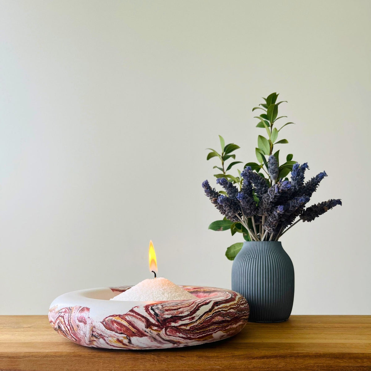 A lit candle in a shallow round dish with a marbled red, white, and pink pattern, placed on a wooden surface next to a ribbed blue-gray vase holding green leaves and purple flowers, against a plain light wall.