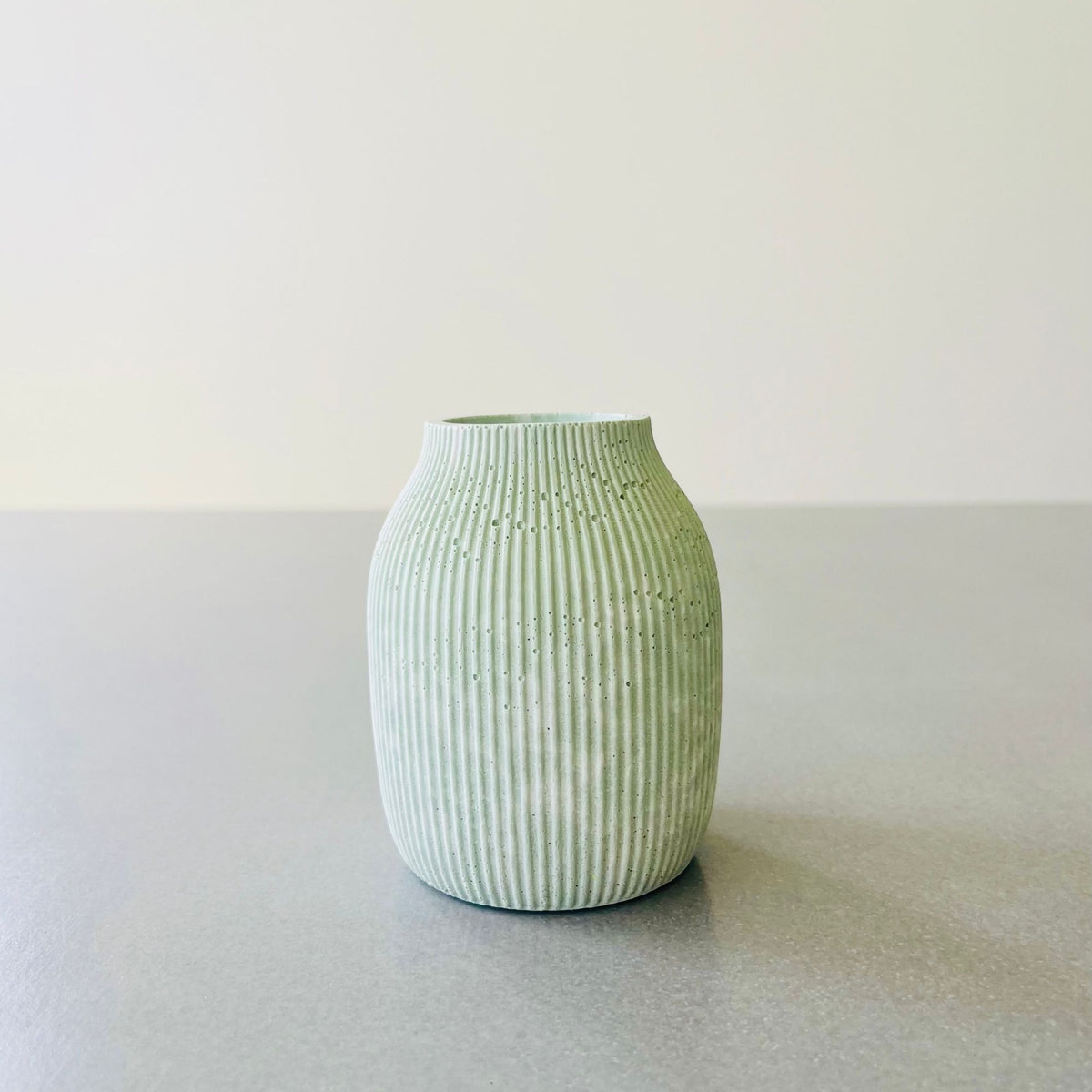 A small ribbed vase in Sea Meadow Marble Concrete finish, standing on a grey surface against a light grey background.